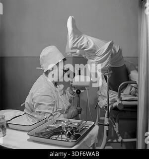 Slaviansk, RSS d'Ukraine, années 1970 Un cliché historique représente une gynécologue féminine concentrée effectuant une colposcopie dans un hôpital soviétique. Le médecin, portant une blouse de coton blanc et une casquette, regarde à travers l'instrument optique pendant un examen du patient. Le patient est placé sur une chaise gynécologique recouverte d'un drap. Un plateau avec des spéculums métalliques stérilisés se trouve au premier plan. Cette authentique photographie d'archives monochrome illustre les normes élevées en matière de soins de santé des femmes et le travail dévoué des spécialistes médicaux dans la paisible région du Donbass. Banque D'Images