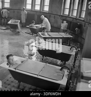 Slaviansk, RSS d'Ukraine, années 1970 Un grand plan historique représente une séance de balnéothérapie dans le sanatorium de la célèbre station balnéaire de Slavkurort. Plusieurs patients masculins se détendent dans des baignoires individuelles remplies d'eau minérale thérapeutique dans un hall carrelé spacieux. Des assistantes médicales en uniforme de coton blanc surveillent la procédure. De grandes fenêtres éclairent la pièce. Cette authentique photographie d'archives monochrome capture l'atmosphère du tourisme de santé soviétique et les pratiques curatives dans la paisible région du Donbass Banque D'Images