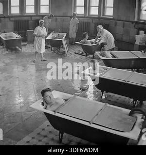 Slaviansk, RSS d'Ukraine, années 1970 Un grand plan historique représente une séance de balnéothérapie dans le sanatorium de la célèbre station balnéaire de Slavkurort. Plusieurs patients masculins se détendent dans des baignoires individuelles remplies d'eau minérale thérapeutique dans un hall carrelé spacieux. Des assistantes médicales en uniforme de coton blanc surveillent la procédure. De grandes fenêtres éclairent la pièce. Cette authentique photographie d'archives monochrome capture l'atmosphère du tourisme de santé soviétique et les pratiques curatives dans la paisible région du Donbass Banque D'Images