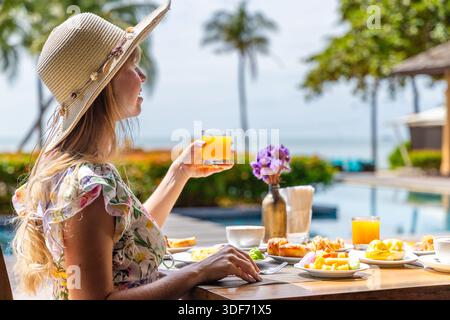 Femme apprécie le petit déjeuner tropical au bord de la piscine à la station, portant un chapeau de soleil, tenant du jus d'orange. Détente, voyage de loisirs, vibrations de vacances dans le plein air serein Banque D'Images