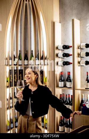 Une femme souriante profite d'un verre de vin tout en étant entourée de bouteilles élégamment agencées dans un élégant espace viticole. Banque D'Images