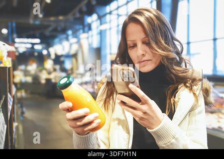 Dans un supermarché lumineux, une jeune femme blonde lit l'étiquette de jus tout en tenant son téléphone, s'assurant qu'elle fait des choix sains pour son alimentation à ce moment-là Banque D'Images