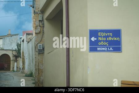 Scène de rue de village grecque traditionnelle avec un panneau directionnel bleu sur un mur pâle, pointant vers Exanti et la route nationale. Banque D'Images
