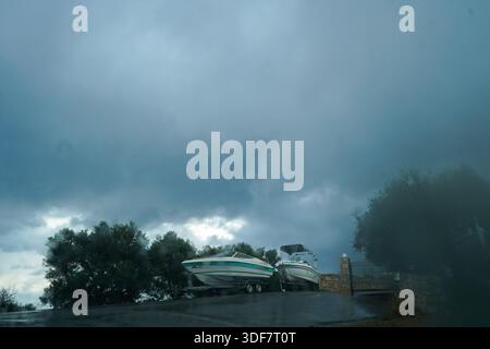Deux bateaux à moteur sur remorques garés à l'extérieur sous un ciel orageux dramatique et sombre. Vue cinématographique à travers un pare-brise couvert de gouttes de pluie en cas de fortes pluies. Banque D'Images