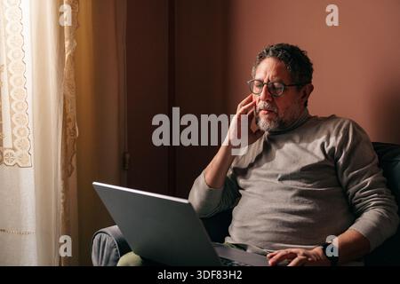 Homme senior travaillant à la maison avec un ordinateur portable près d'une fenêtre, réfléchi et stressé tout en analysant des informations à l'écran dans une lumière naturelle chaude. Banque D'Images