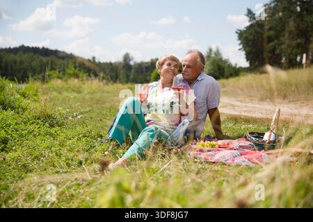 charmant couple de personnes âgées au lac ayant un pique-nique, faisant un toast sur la rive du lac un jour d'été. Staycations, voyage hyper-local, sortie en famille, escapade. || autorisation du modèle Banque D'Images