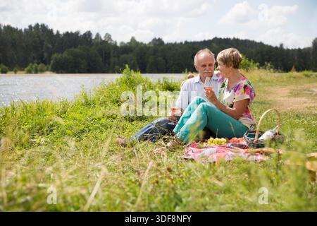 charmant couple de personnes âgées au lac ayant un pique-nique, faisant un toast sur la rive du lac un jour d'été. Staycations, voyage hyper-local, sortie en famille, escapade. || autorisation du modèle Banque D'Images