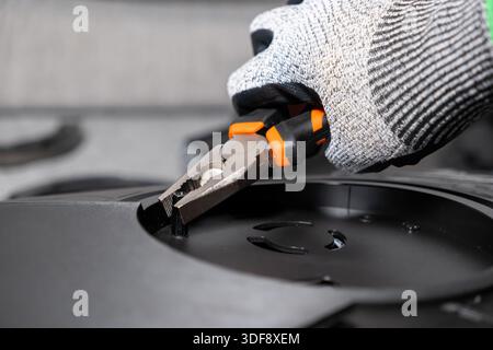 Gros plan de la main d'un travailleur portant des gants de protection, tenant une pince pointue pour manipuler délicatement une petite pièce en plastique noir sur un mécanicien complexe Banque D'Images