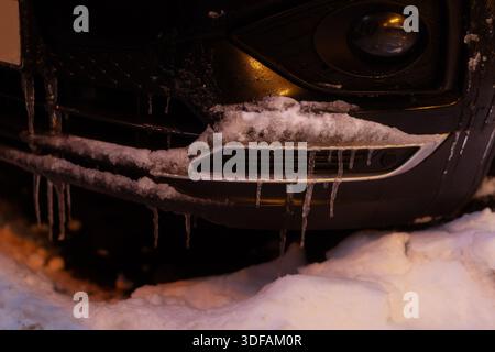Gros plan d'une calandre et d'un pare-chocs sombres de voiture fortement recouverts de givre et de glaçons suspendus, illustrant les effets des températures hivernales extrêmes. Banque D'Images