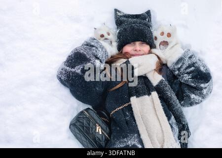 portrait d'une jeune fille preteen joyeuse portant un manteau noir confortable, une écharpe blanche et un bonnet beanie mignon aux oreilles de chat, couché confortablement sur la neige fraîche. Authen Banque D'Images