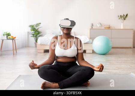 Femme noire de taille plus dans un casque VR, assise dans une posture lotus, méditant ou pratiquant le yoga à l'intérieur Banque D'Images