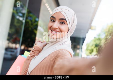 Joyeuse heureuse jolie blogueuse acheteuse arabe millénaire en hijab avec beaucoup de sacs fait selfie sur l'appareil Banque D'Images