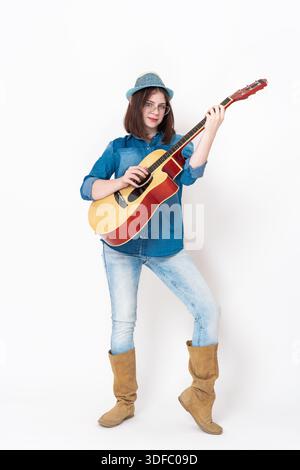 Joueur de guitare femme jouant de la guitare acoustique et regardant la caméra, debout sur toute la longueur. Jeune femme de Caucase, portant des lunettes, habillée en str Banque D'Images