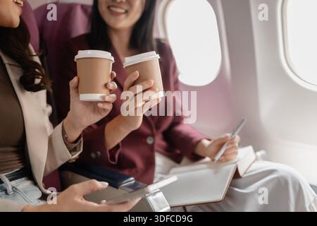 Deux femmes s'assoient ensemble dans un avion, souriant et tenant des tasses de café par la fenêtre, exprimant une interaction amicale, des moments de voyage partagés, et un Banque D'Images
