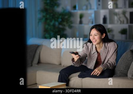 Femme asiatique heureuse utilisant la télécommande regardant la télévision dans la nuit à la maison Banque D'Images