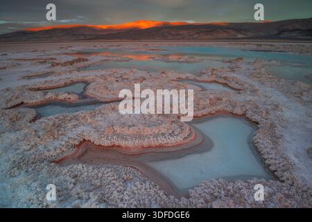 Formations de croûte salée et bassins peu profonds dans le bassin de Badwater, dans la vallée de la mort, au coucher du soleil. Montagnes en arrière-plan avec alpenglow orange. Banque D'Images
