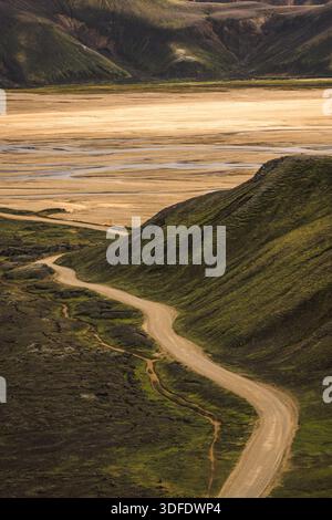 Vue sur un chemin de terre sinueux qui traverse des collines verdoyantes menant à une vaste plaine ensoleillée à Landmannalaugar, les Hautes terres centrales, en Islande. Banque D'Images