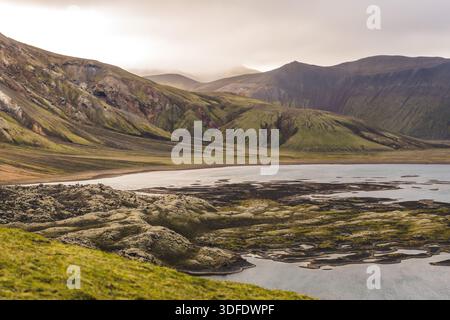 Vue sur les champs de lave accidentés couverts de mousse et les eaux scintillantes du lac sous les montagnes imposantes dans les Highlands islandais, Landmannalaugar, le C. Banque D'Images