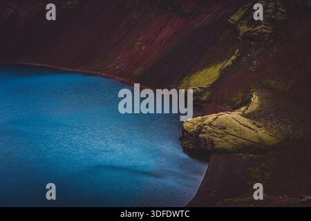 Vue sur l'eau bleue sereine nichée contre le contraste frappant du paysage volcanique rouge et noir, avec des taches de mousse verte, Landmannalaugar, Banque D'Images