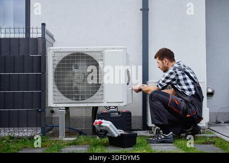 Technicien effectuant l'entretien de la pompe à chaleur extérieure près de l'extérieur du bâtiment résidentiel. Homme installant la pompe à chaleur de source d'air, à l'aide d'outils. Conc Banque D'Images