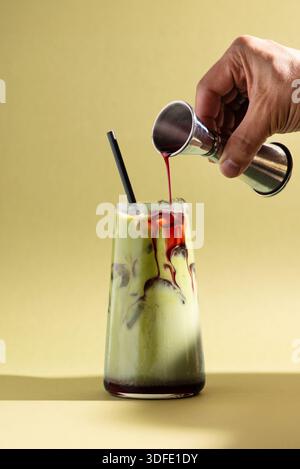 Boisson matcha à la framboise glacée avec du sirop rouge versé d'un jigger dans un verre transparent. Boisson colorée en couches. Banque D'Images