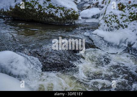 Un ruisseau coule à travers des zones rocheuses et coule rapidement. La neige et la glace se trouvent sur les bords. La zone environnante est calme et montre le paysage hivernal. Banque D'Images