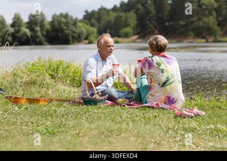 charmant couple de personnes âgées au lac ayant un pique-nique, faisant un toast sur la rive du lac un jour d'été. Staycations, voyage hyper-local, sortie en famille, escapade. || autorisation du modèle Banque D'Images