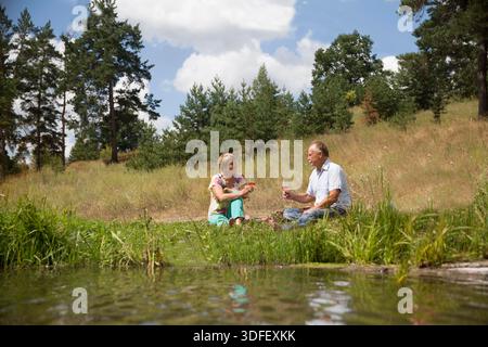 charmant couple de personnes âgées au lac ayant un pique-nique, faisant un toast sur la rive du lac un jour d'été. Staycations, voyage hyper-local, sortie en famille, escapade. || autorisation du modèle Banque D'Images