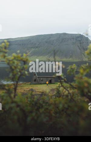 Vue d'une figure solitaire se dresse devant une structure en bois abandonnée de maison A par les eaux tranquilles d'un fjord entouré de collines verdoyantes, Westfjord Banque D'Images