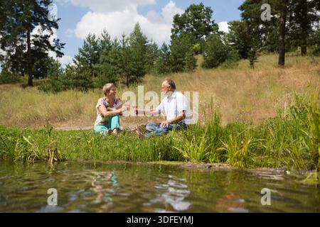 charmant couple de personnes âgées au lac ayant un pique-nique, faisant un toast sur la rive du lac un jour d'été. Staycations, voyage hyper-local, sortie en famille, escapade. || autorisation du modèle Banque D'Images