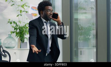 Homme d'affaires afro-américain en colère criant sur le téléphone portable dans l'homme d'affaires de bureau moderne jetant des documents dans la frustration regardant hors du mâle de fenêtre Banque D'Images