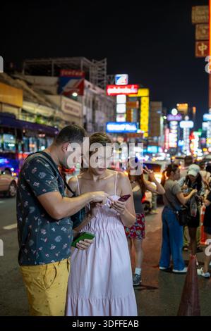 Bangkok, Thaïlande - 11.11.2025, deux touristes se tenant sur la route de Yaowarat à Bangkok Chinatown, regardant un téléphone portable, entourés de néons lumineux sig Banque D'Images