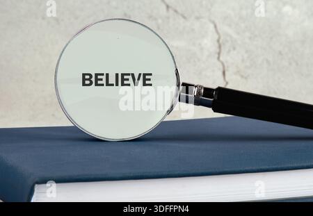 Une loupe affiche le mot croire. Il repose sur un livre bleu fermé. Le livre est sur une surface blanche devant un BA à motifs gris et blanc Banque D'Images