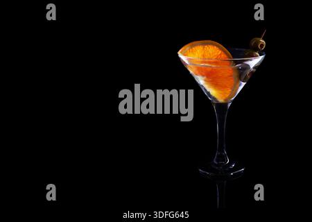 Cocktail de martini clair dans un verre, garni d'une tranche d'orange éclatante et d'olives vertes sur un fond noir Banque D'Images