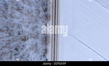 Scène aérienne hivernale à Nowy Lubiel, Pologne. Une route labourée est verticale, divisant une forêt sans feuilles et un champ balayé par le vent avec de faibles traces d'animaux sous s. Banque D'Images