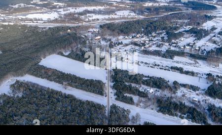 Vue aérienne de Nowy Lubiel, Pologne, montrant la neige sur les champs et les pinèdes. Une route rurale droite mène au village, avec des fermes, un toit rouge près de l'arbre Banque D'Images