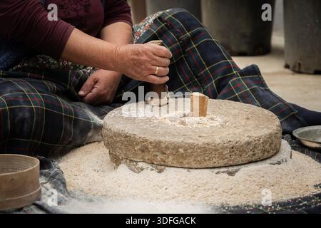 Femme âgée broyant du blé entier en farine avec un moulin à main en pierre traditionnel, reflétant l'agriculture ancienne et la préparation naturelle des aliments Banque D'Images