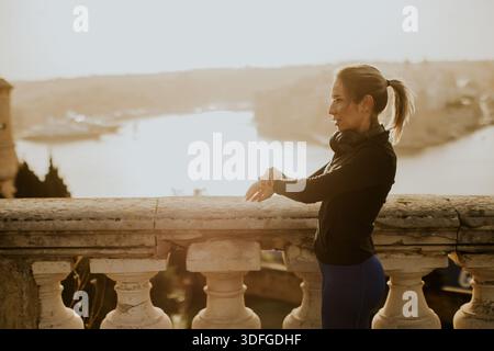 Une femme en tenue d'entraînement s'étire alors que le soleil se couche sur la magnifique côte maltaise. La scène tranquille améliore son parcours de remise en forme. Banque D'Images