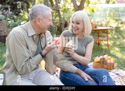 Rire les conjoints âgés assis sur l'herbe de champ et manger des fruits, parler et sourire, se reposer à l'extérieur dans leur jardin Banque D'Images