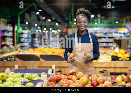 Femme afro-américaine portant un tablier et des gants, souriant tout en tenant des pommes fraîches dans la section des grands fruits et légumes d'une épicerie moderne, symboli Banque D'Images