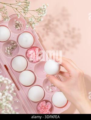 Placer à la main l'œuf de Pâques blanc dans un conteneur rose avec des fleurs sur fond rose. Concept des traditions de Pâques, Palm Sanday, printemps renouveau et Eleg Banque D'Images