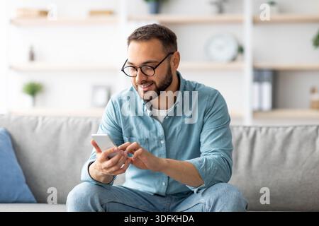 Jeune garçon positif en lunettes assis sur un canapé avec téléphone mobile et textant à son ami, à l'intérieur Banque D'Images