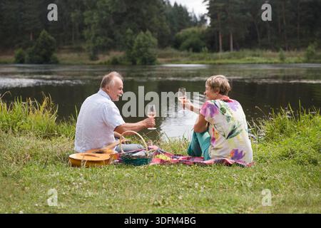 charmant couple de personnes âgées au lac ayant un pique-nique, faisant un toast sur la rive du lac un jour d'été. Staycations, voyage hyper-local, sortie en famille, escapade. || autorisation du modèle Banque D'Images