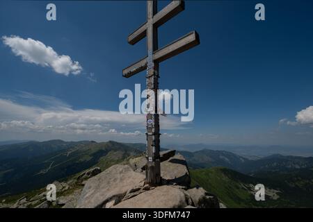 Vue d'une croix en bois altérée debout fièrement au sommet d'un pic rocheux, perçant le ciel bleu clair au-dessus des chaînes de montagnes lointaines, Nizke Tatry, Zilin Banque D'Images