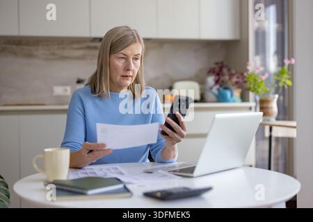 Femme senior à la table de cuisine examinant les factures et la paperasse du ménage, en utilisant un ordinateur portable et un smartphone pour les services bancaires en ligne, la budgétisation et la planification de la retraite avec une concentration ciblée Banque D'Images