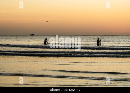 Vue du coucher de soleil doré projetant de longues ombres sur la plage, avec des silhouettes de gens pataugant dans les vagues douces, Kuta Beach, Bali, Indonésie. Banque D'Images