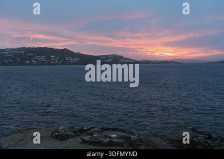 Vue sur la mer tranquille et bleu foncé reflétant le soleil couchant vibrant et rayé rose sur les collines lointaines, avec un cercle de pierre au premier plan, Mykono Banque D'Images