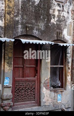 Vue d'une porte rouge altérée avec des sculptures en bois complexes se dresse à côté d'une fenêtre barrée contre un mur vieilli et texturé, Lahore, Punjab, Pakistan. Banque D'Images