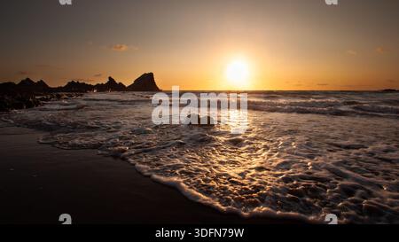 Le soleil se couche sur l'océan, projetant une lueur chaude sur les vagues alors qu'elles s'écrasent contre la rive rocheuse. Banque D'Images