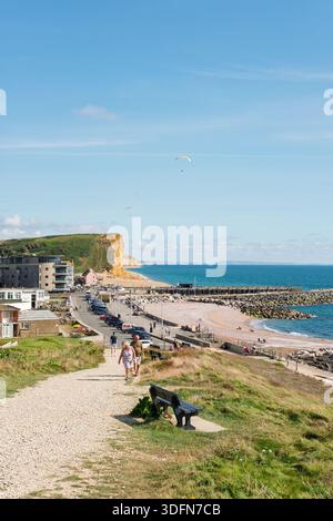 West Bay Dorset 9-14-2023 Un couple se promène le long d'un sentier côtier pittoresque avec des parapentes dans le ciel au-dessus de la plage et du port. Banque D'Images